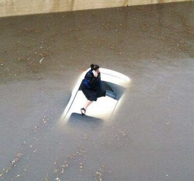 A photograph of a woman stranded on top of a car hood in the middle of a flood. She appears to be on her phone making a call