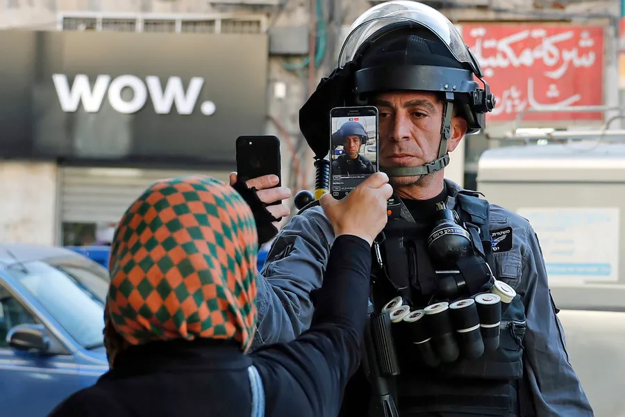 A photograph of a police officer in riot gear and a woman in a head scarf who appear to be standing off. Each person is pointing their smart phone at the other, presumably recording one another