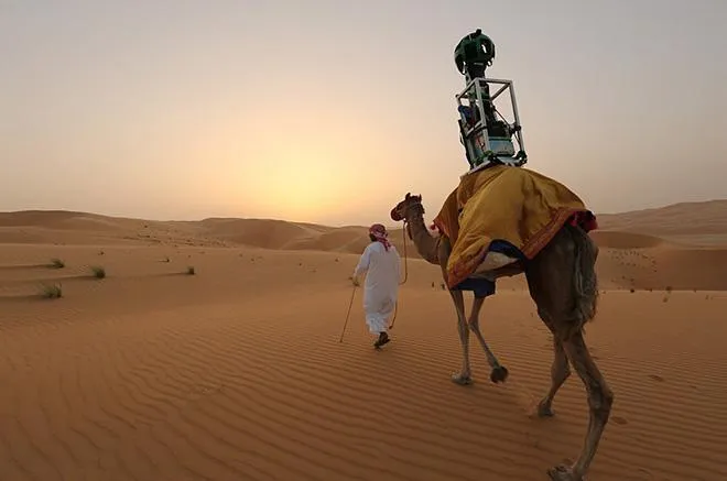 A photograph of a man and a camel in what appears to be the Saharan desert. The camel has an apparatus strapped atop its back, which looks like a Google Maps photo sphere, for recording the surrounding environment